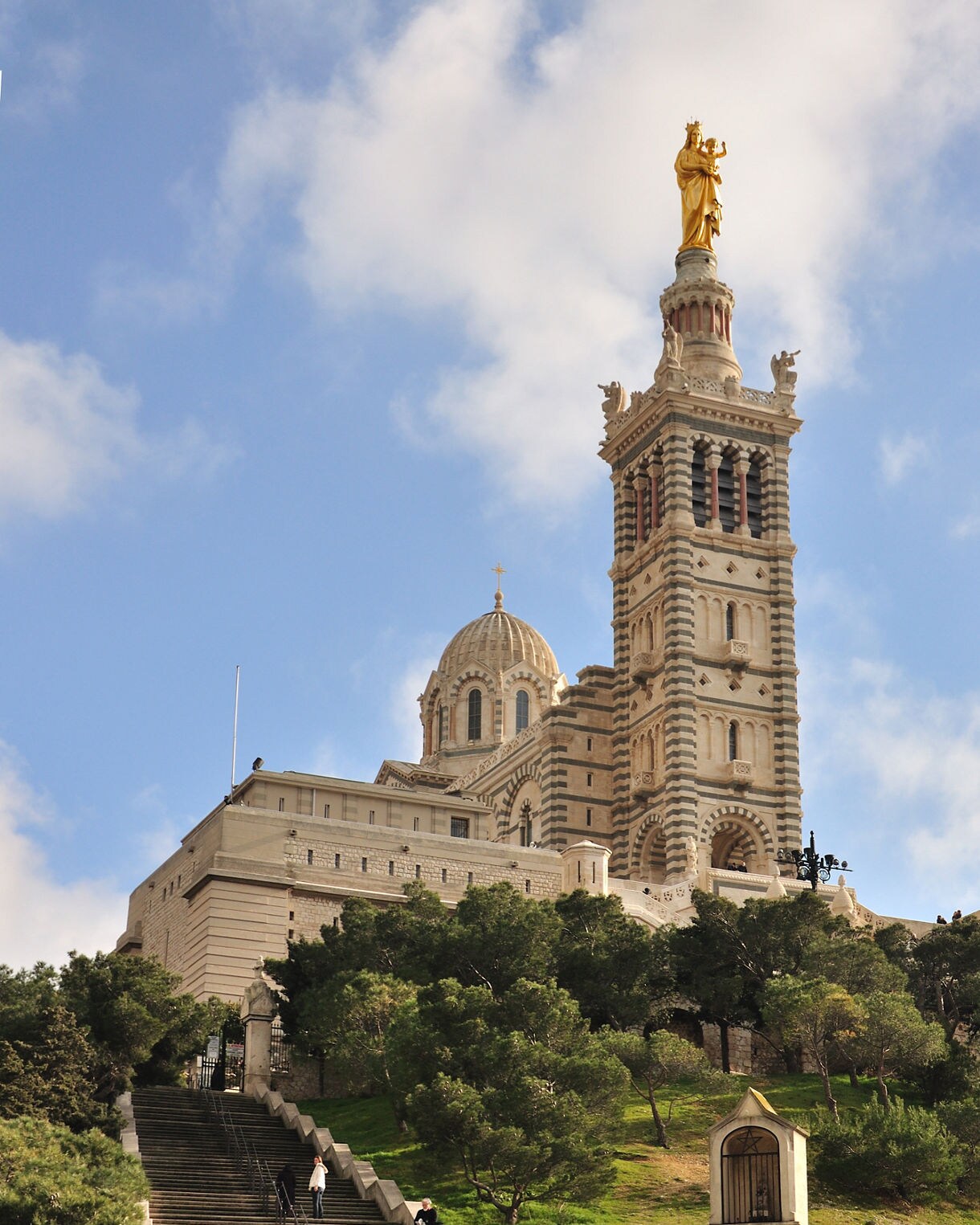 Exterior view of Notre-Dame de la Garde in Marseille, with its striped stone façade, domed roof and golden statue of the Virgin Mary atop the bell tower against a blue sky.