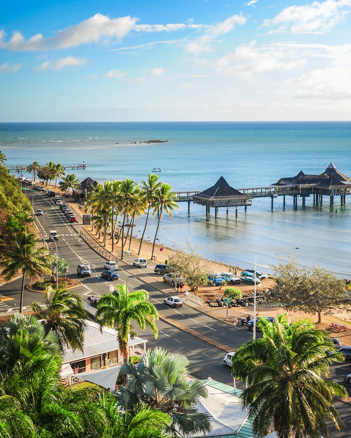 Elevated view of a coastal road in Nouméa bordered by palm trees, with a sandy beach and overwater pavilions extending into calm turquoise water under a bright blue sky.
