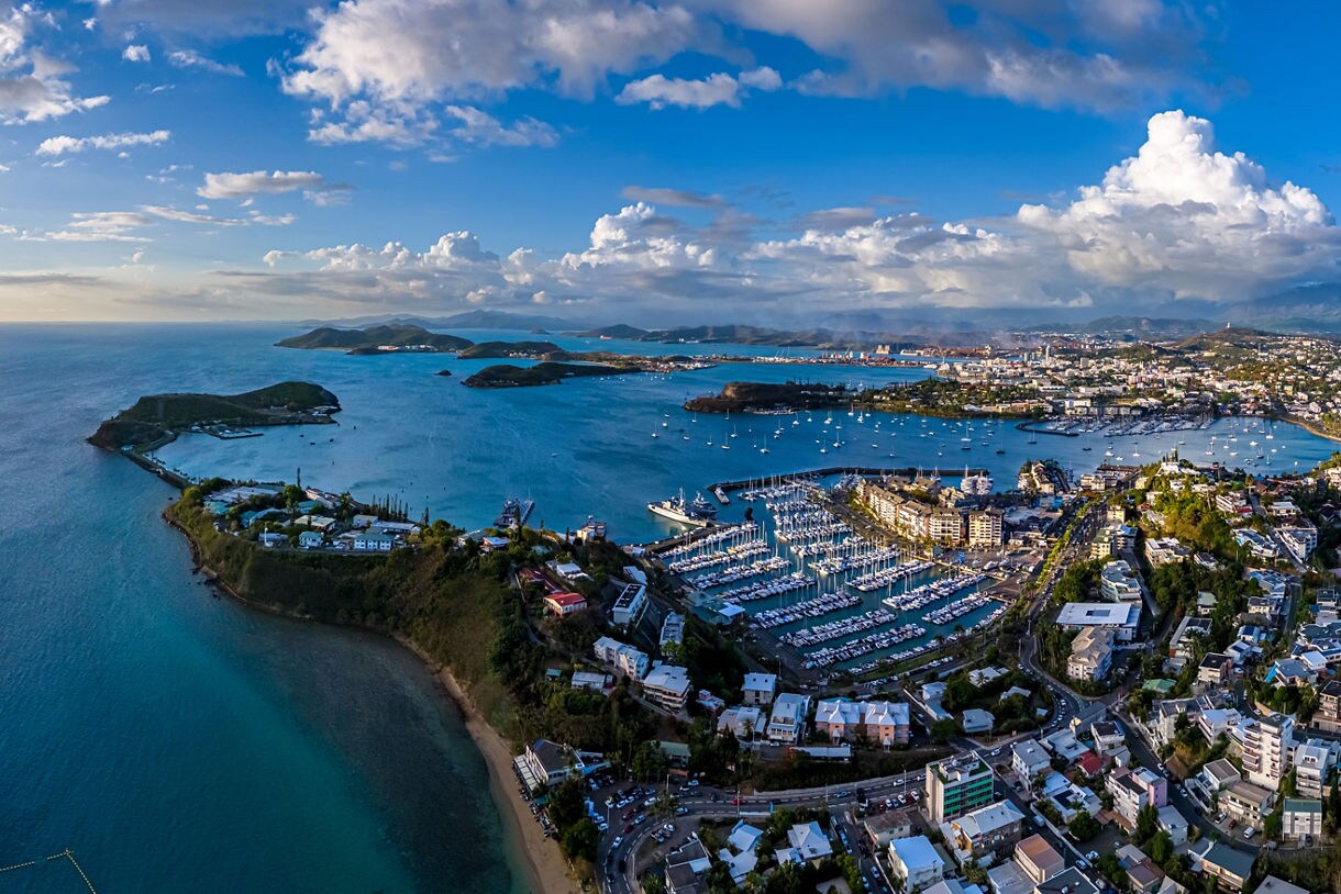 Aerial view of Nouméa showing marinas filled with sailboats, turquoise bays, small islands and a sprawling city backed by green hills under a bright sky.Aerial view of Nouméa showing marinas filled with sailboats, turquoise bays, small islands and a sprawling city backed by green hills under a bright sky.
