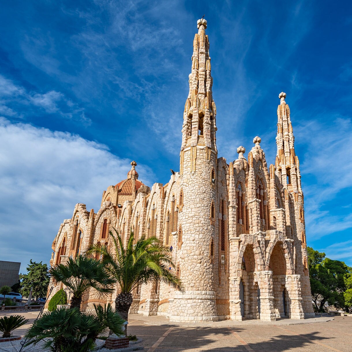 Stone sanctuary in Novelda with tall ornate towers, arched entrances and textured walls set beneath a bright blue sky with scattered clouds.