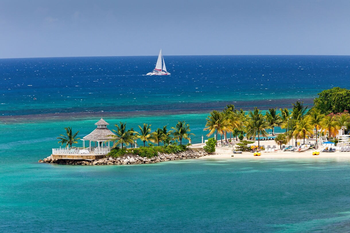 Tropical beach in Ocho Rios, Jamaica, with palm trees, lounge chairs and a sailboat on the vivid turquoise sea.
