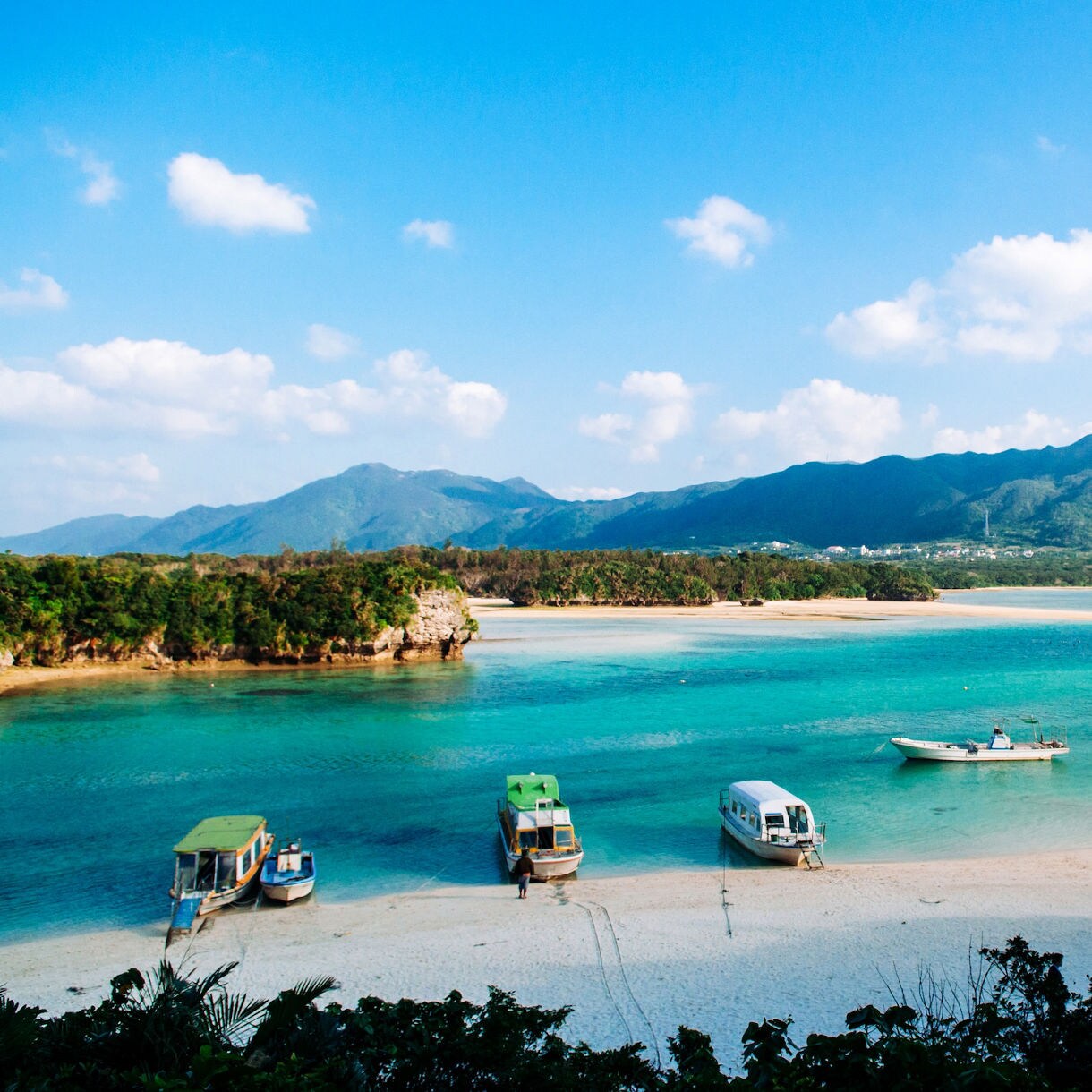 A secluded sandy cove in Okinawa, Japan, with crystal-clear shallow water lapping the shore, dramatic limestone rocks draped in lush tropical vegetation and distant blue islands visible across the open sea under a brilliant blue sky.