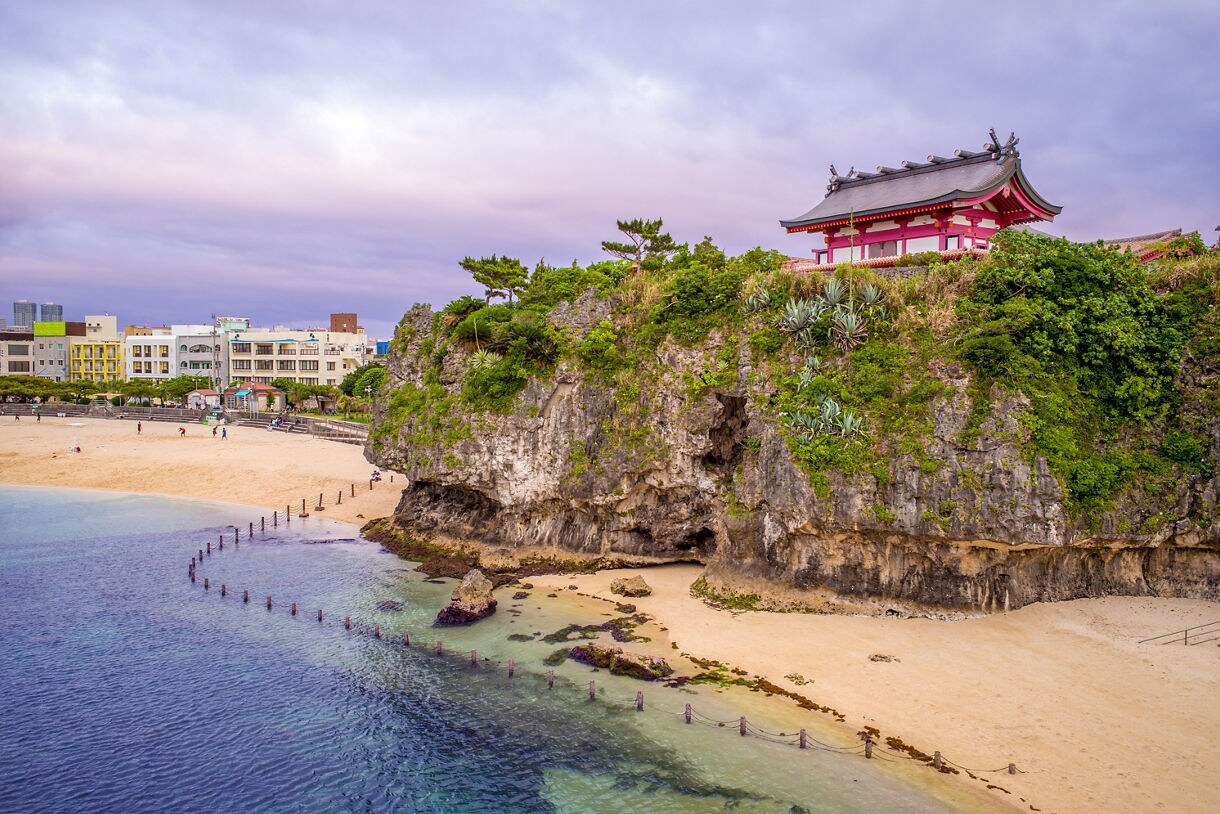 Traditional Okinawan shrine perched atop a rocky cliff above a sandy beach, with clear blue water, coastal greenery and nearby city buildings in the background.