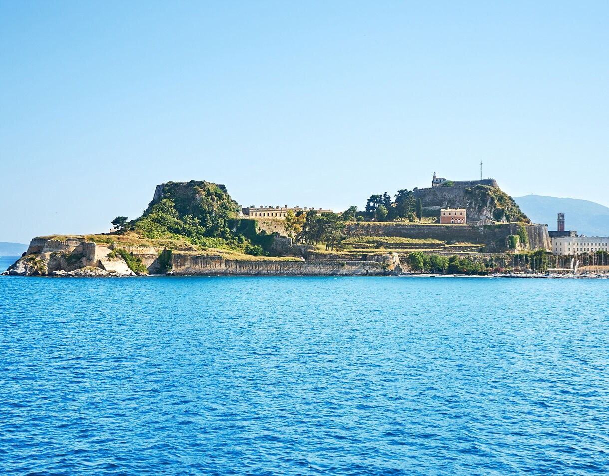 The Old Fortress of Corfu, a historic Venetian fort built on a rocky peninsula, viewed from the sea against a backdrop of blue water and mountains.