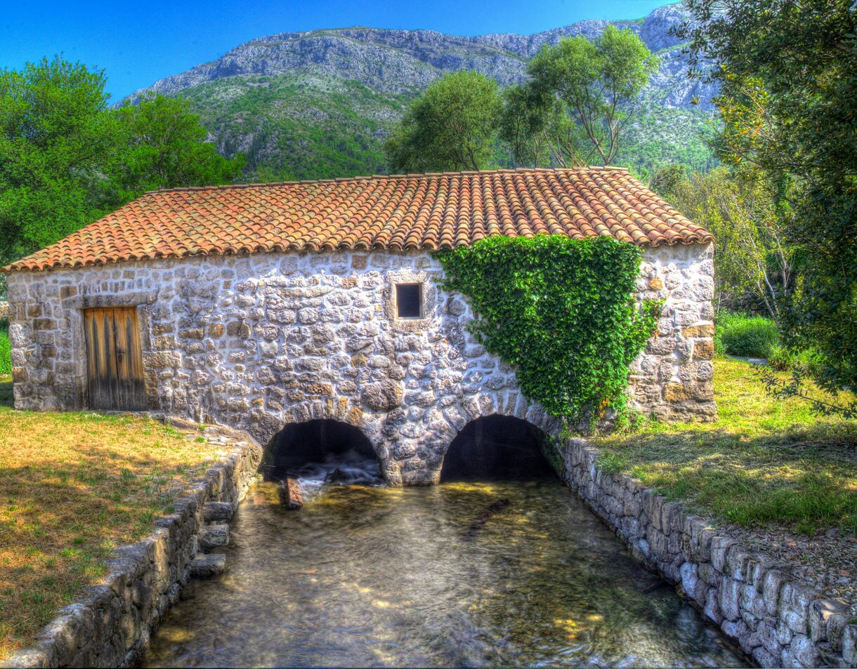 Stone-built Old Mill in Ljuta with a red-tiled roof, ivy climbing the wall and water running beneath its arches against a backdrop of green trees and mountains.