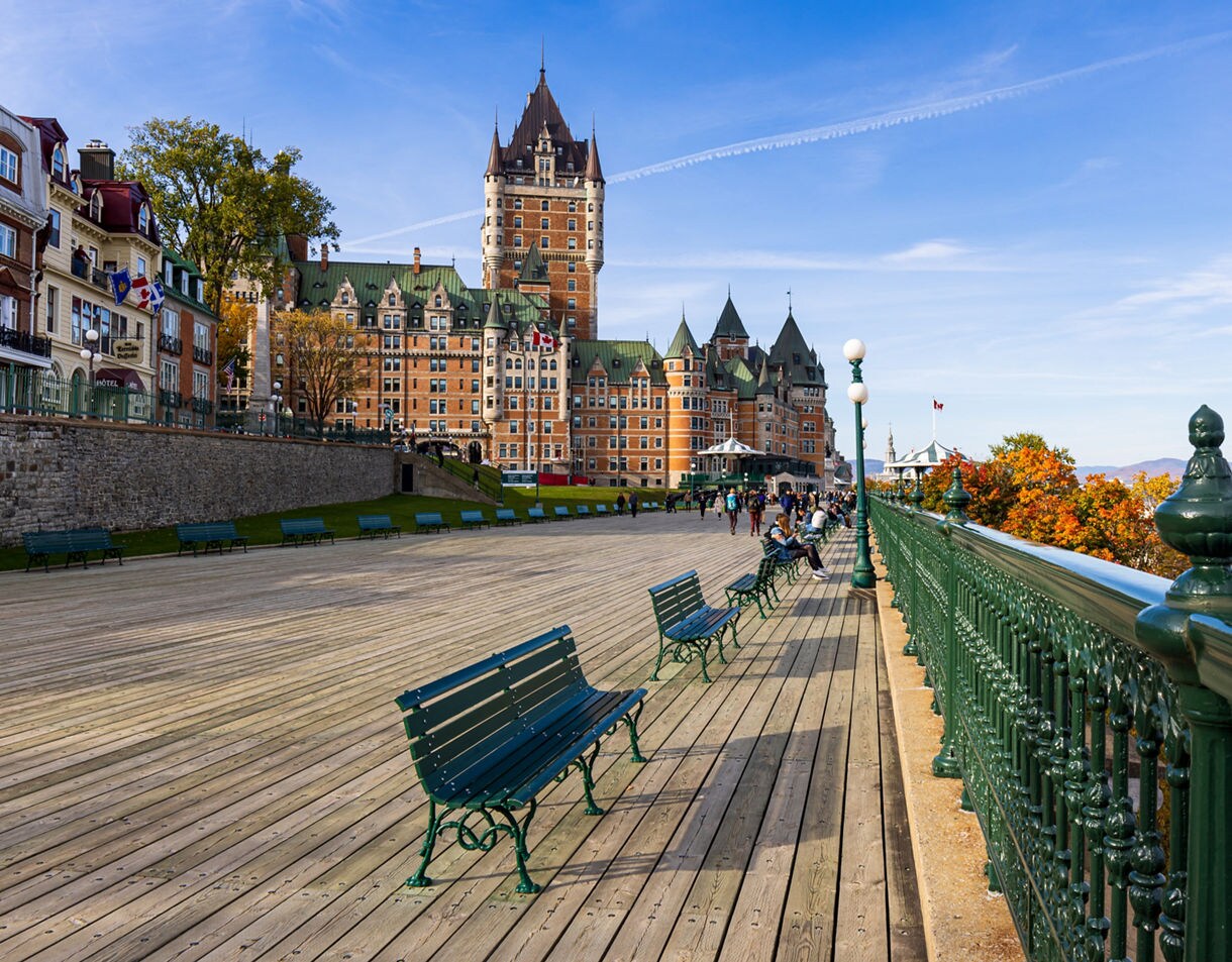 View of Québec City’s Dufferin Terrace with green benches, a decorative railing and the Château Frontenac towering behind historic buildings under a clear blue sky.