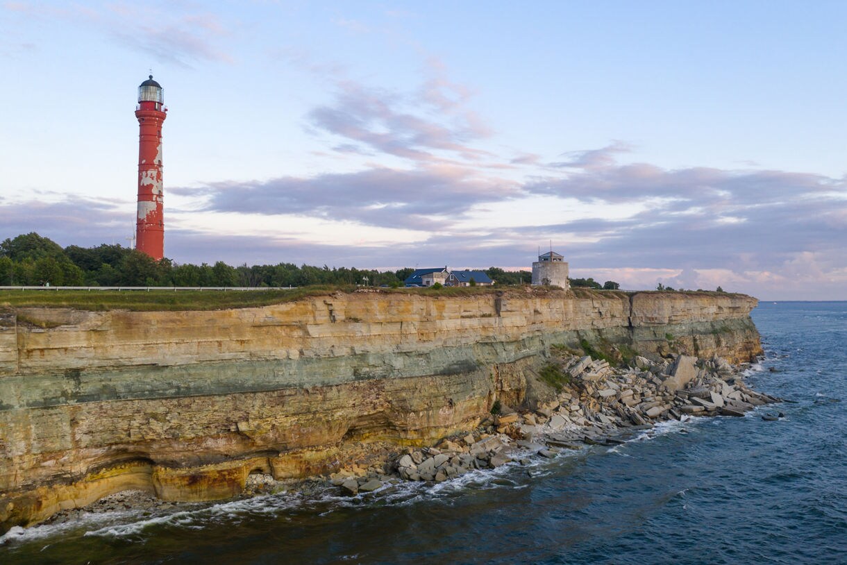 View of Pakri Lighthouse near Tallinn, Estonia, a tall red tower perched on rocky limestone cliffs overlooking the Baltic Sea under a pastel evening sky.