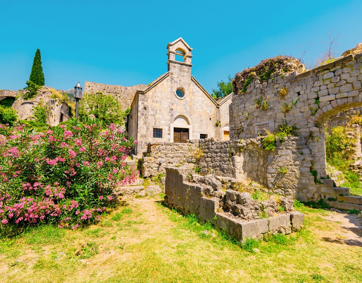 Stone church and ruined walls surrounded by bright pink oleander and green grass in Stari Bar, Montenegro, under a clear blue sky.