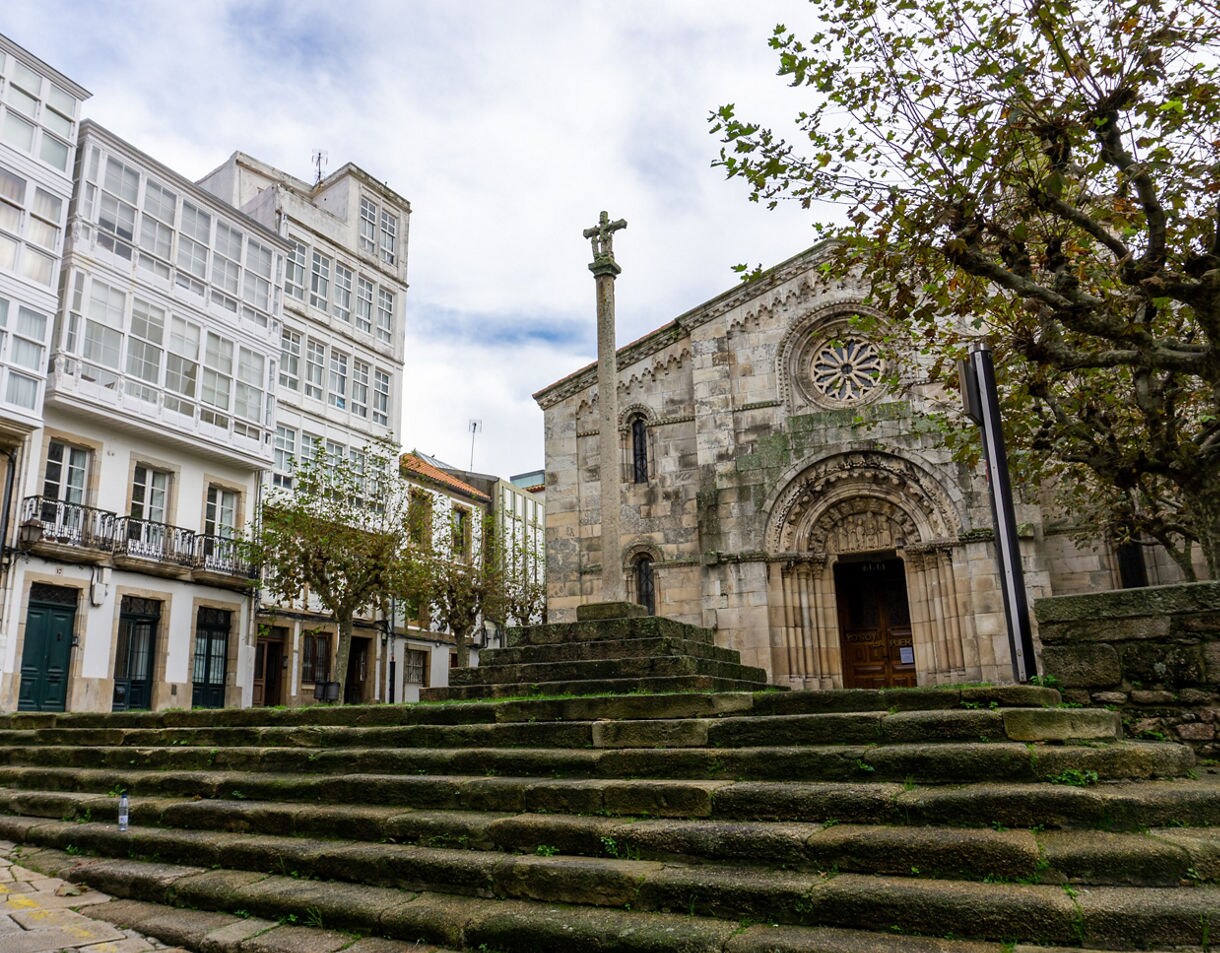 Small historic plaza in A Coruña with worn stone steps leading to a Romanesque church, alongside traditional Galician buildings with white enclosed balconies.
