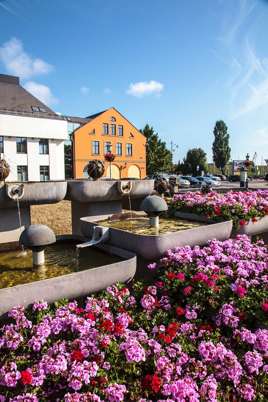 Flower-filled square in Klaipėda’s old town with fountains in the foreground and an orange historic building behind.