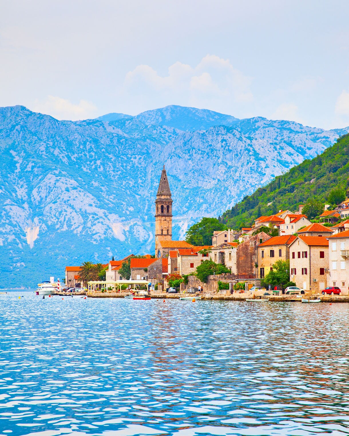 Waterfront view of Montenegro’s old town with red-roofed stone buildings and a tall church bell tower, backed by rugged blue mountains.