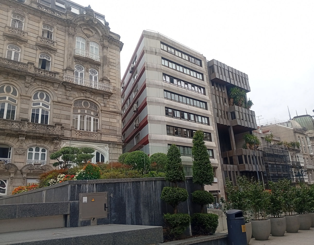 A city street scene showing a historic ornate stone building next to a modern multi-story concrete structure with geometric shapes and greenery on balconies, with trimmed bushes and planters in the foreground.