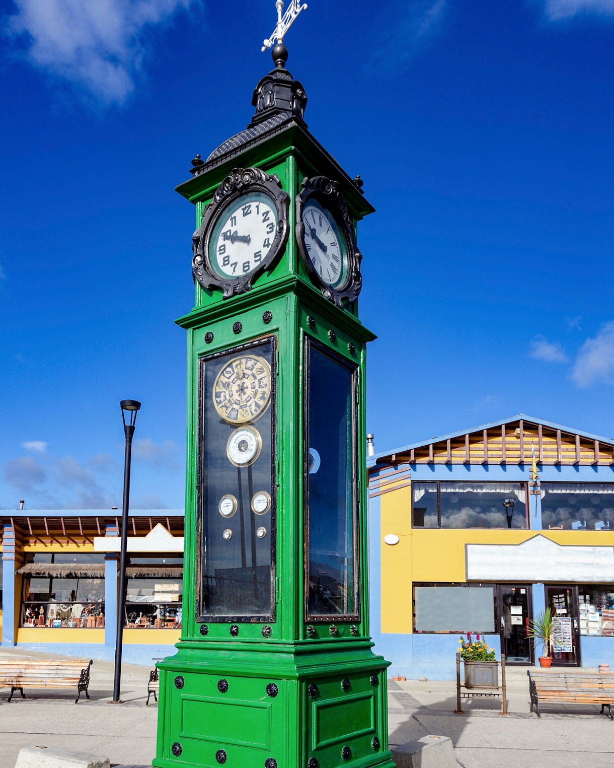 Tall green clock tower with multiple dials and black trim standing in a sunny plaza, with bright blue sky and colorful buildings in the background.