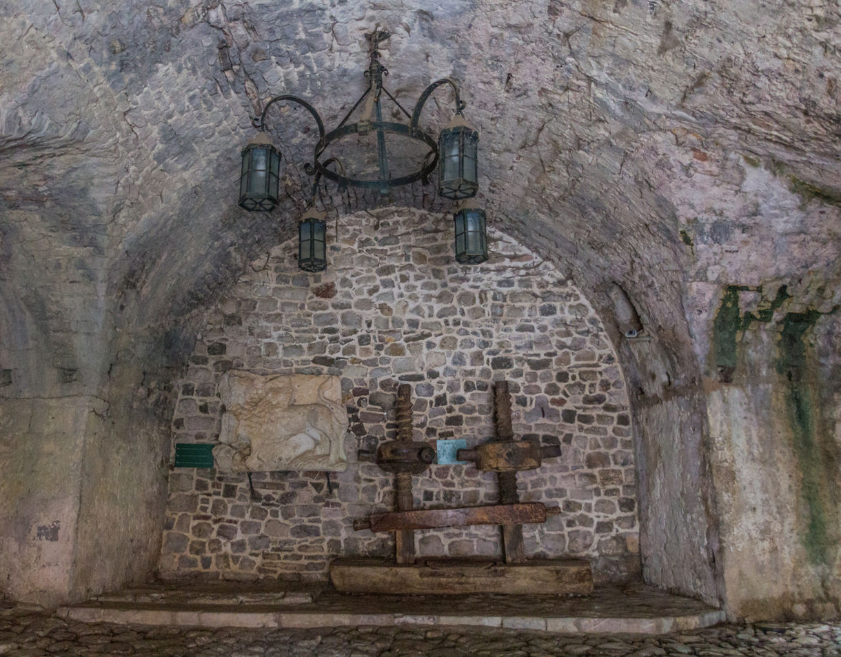 A stone-walled vaulted cellar featuring an antique wooden olive press and a rustic chandelier hanging from the ceiling.