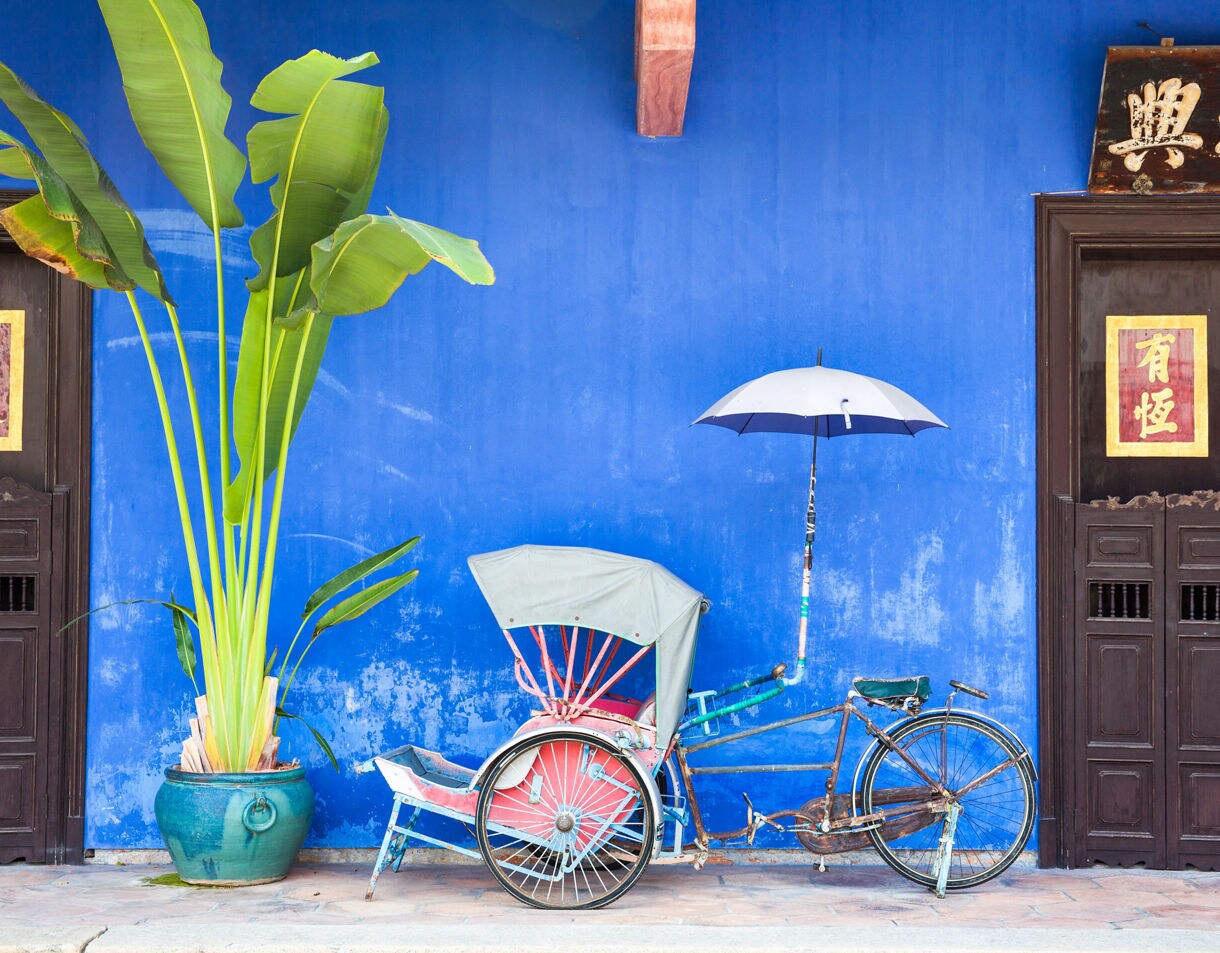 Old rickshaw tricycle with umbrella parked by a bright blue wall and potted plant outside the Cheong Fatt Tze Mansion in Penang.