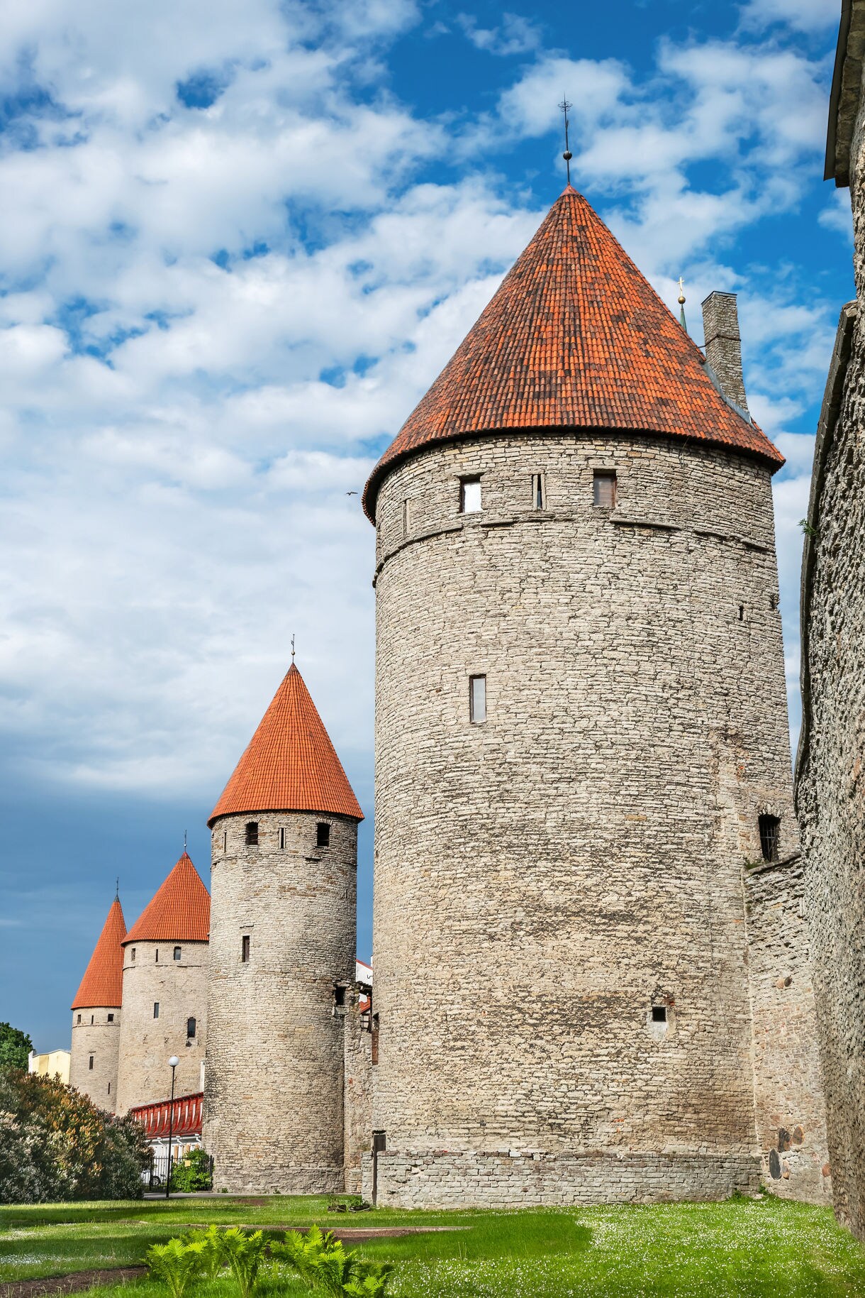 Tall stone towers with red-tiled conical roofs forming part of Tallinn’s historic city wall, set against a partly cloudy blue sky.