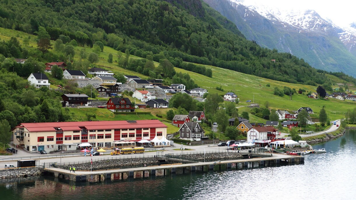The port of Olden, Norway, with a dockside terminal, colorful houses and green hills rising to snow-dusted mountains in the background.