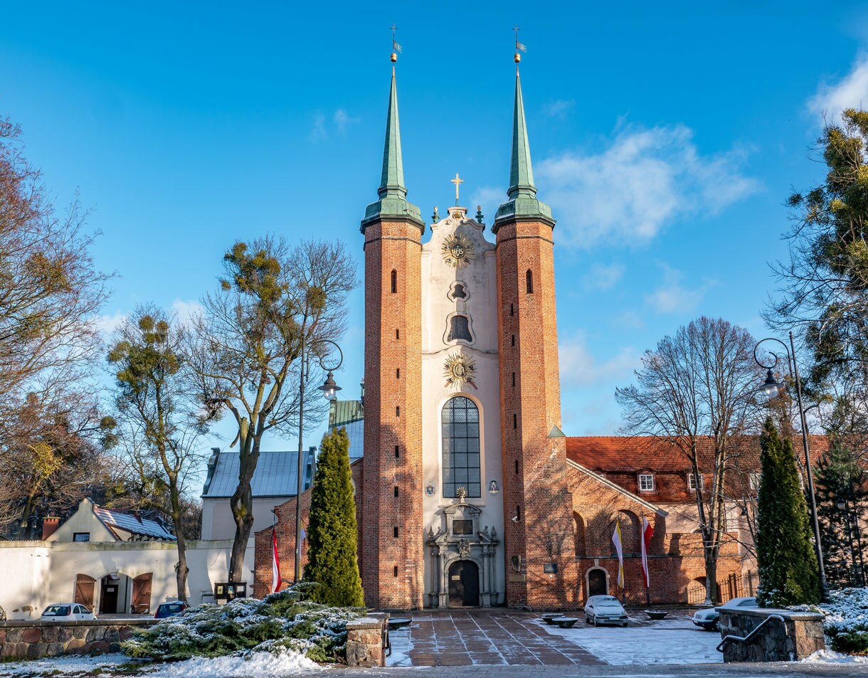 Front view of Oliwa Cathedral in Gdańsk, Poland, featuring two tall spires, brick façade and baroque details framed by trees and a light dusting of snow.