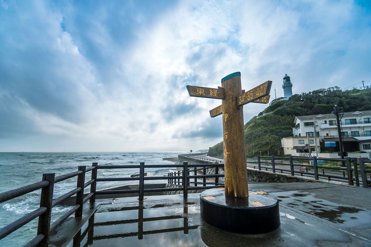 Wooden signpost with Japanese characters standing on a wet coastal promenade above choppy gray waves, with a lighthouse perched on a green hill and seaside houses lining the road in the background.