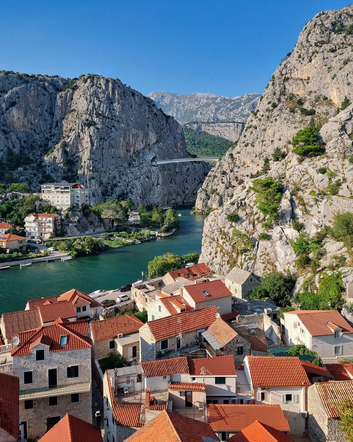 Red-roofed stone houses in Omiš beside the green Cetina River, framed by steep rocky cliffs and a clear blue sky.