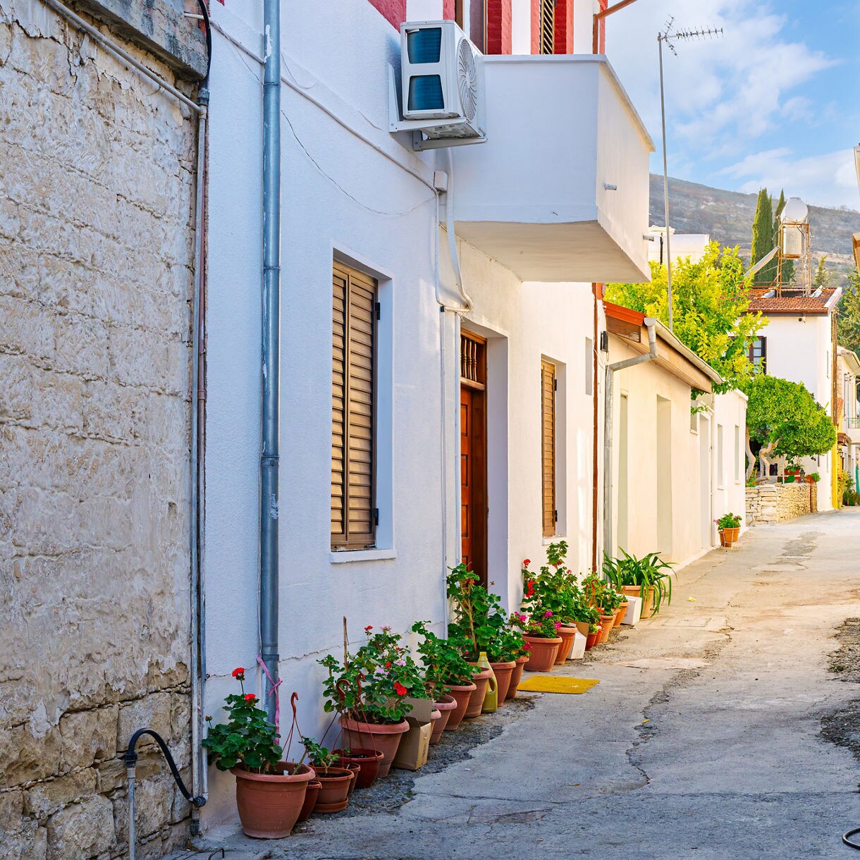 Narrow stone alley in Omodos Village lined with whitewashed homes, potted flowers and rustic balconies, leading toward a sunlit courtyard framed by trees and distant hills.
