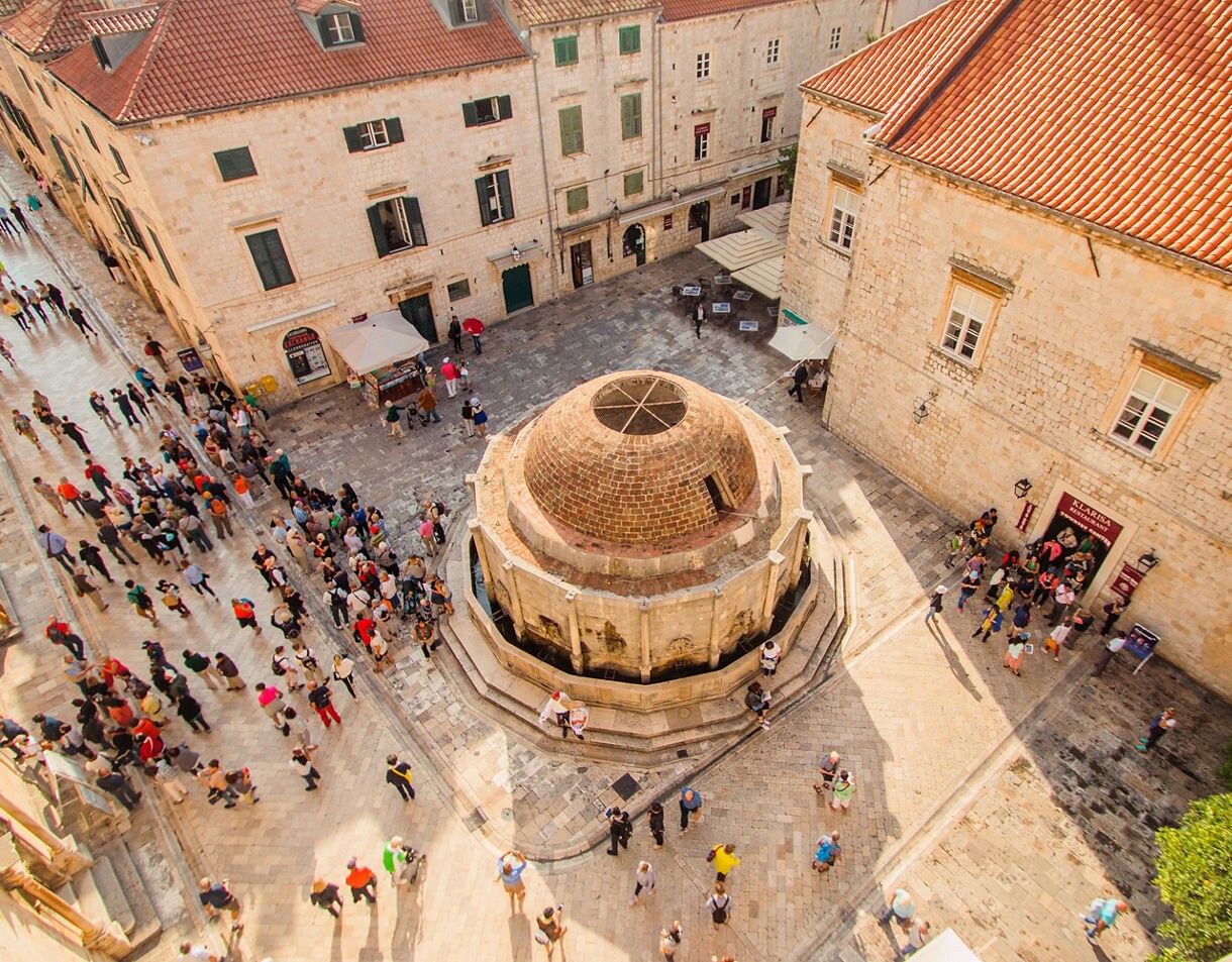 Aerial view of Onofrio’s Fountain in Dubrovnik’s Old Town, surrounded by crowds of visitors in a stone-paved square lined with historic buildings.