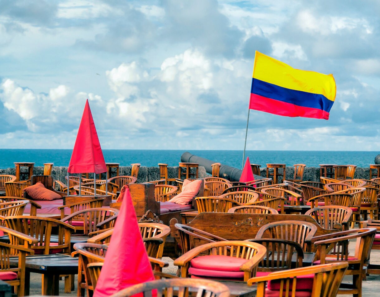 Outdoor café with wooden chairs and red cushions overlooking the sea in Cartagena, featuring the Colombian flag flying above the terrace.