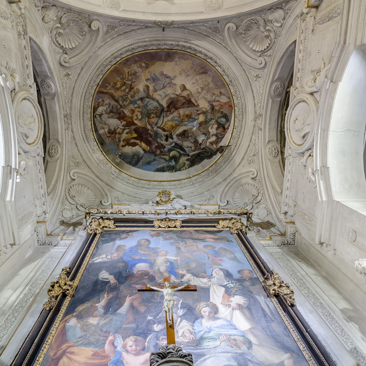Interior of a richly decorated chapel with a large painted fresco on the ceiling and an elaborate framed religious painting with a crucifix at the center.