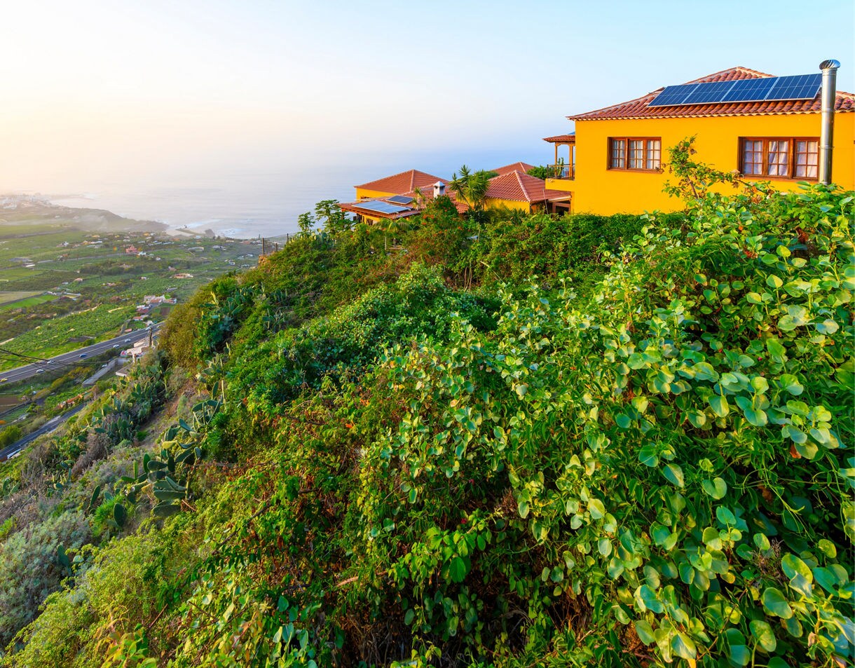 View of a bright yellow hillside home overlooking the green Orotava Valley in Tenerife, with the Atlantic Ocean and coastal town in the distance at sunset.