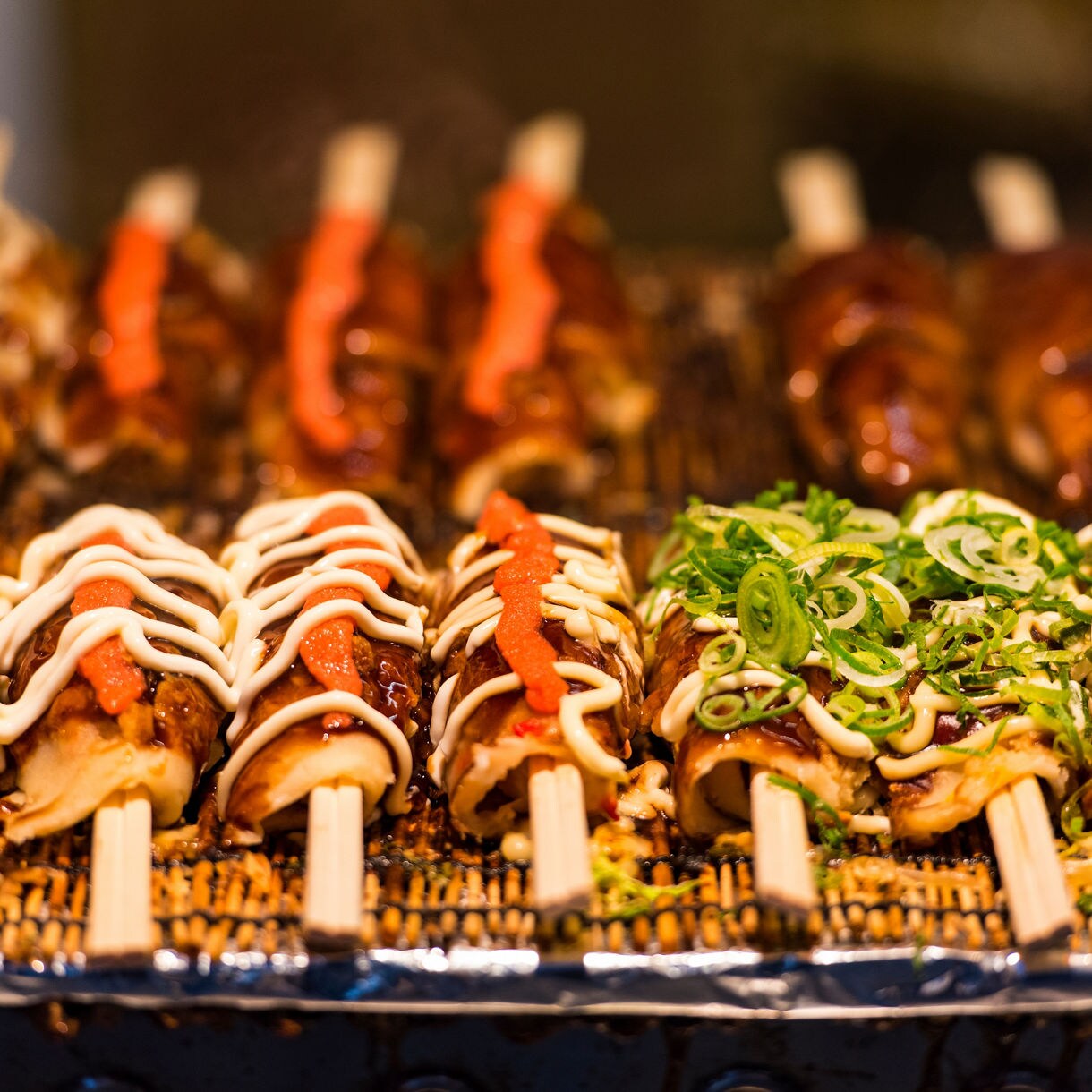 Rows of rolled okonomiyaki topped with mayonnaise, spicy sauce and chopped scallions, displayed on sticks at a street food stall.