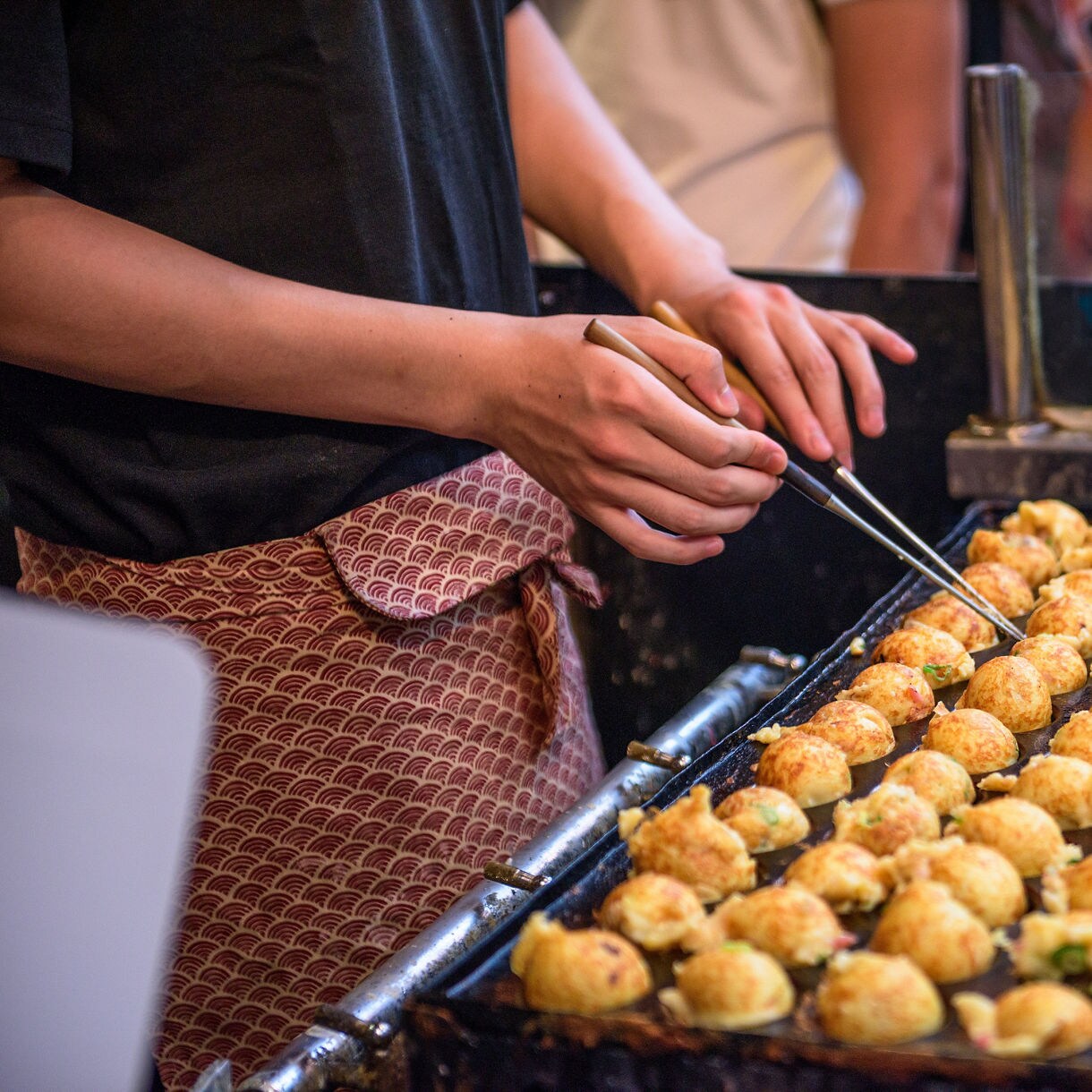 A street vendor in a patterned apron using metal skewers to turn rows of golden takoyaki balls on a large iron griddle at an Osaka street food stall.