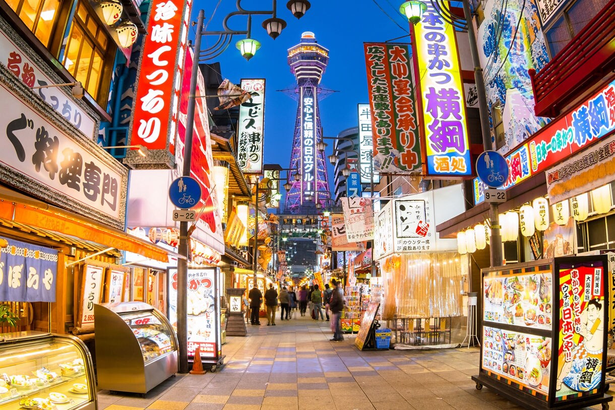 Nighttime street scene in Osaka’s Shinsekai district with glowing restaurant signs, busy pedestrian walkways and Tsutenkaku Tower rising in the background.