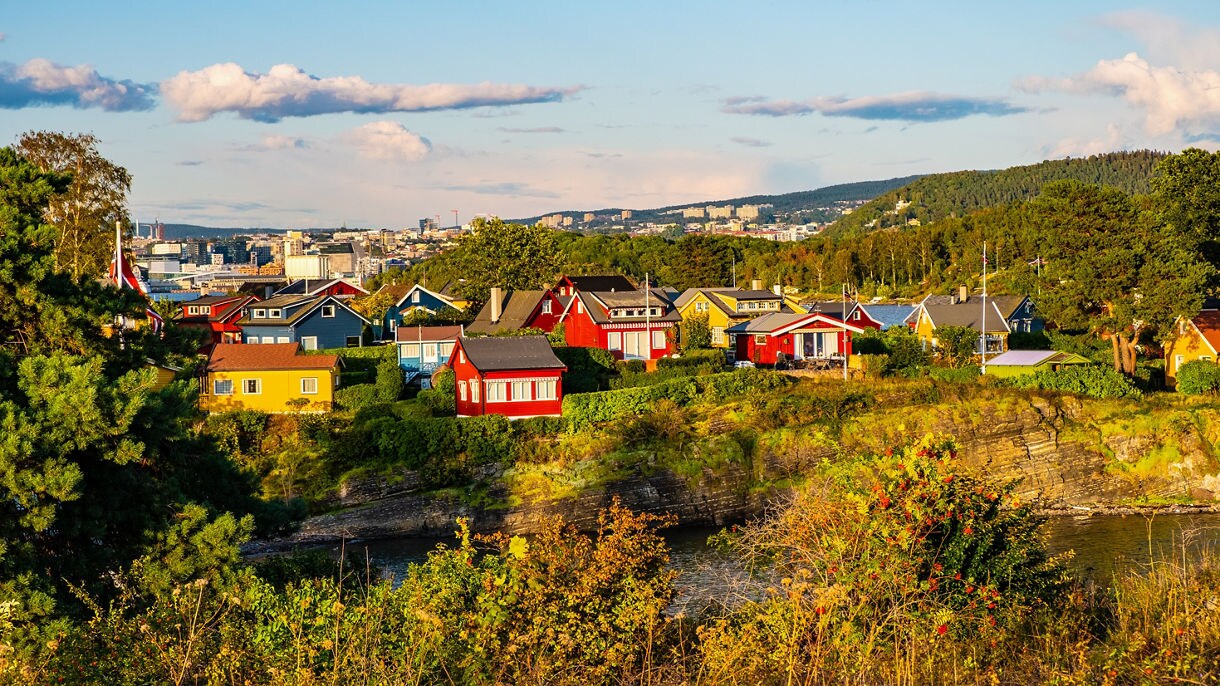 Bright red, yellow and blue houses on a green hillside above Oslo’s harbor with the modern cityscape in the background.