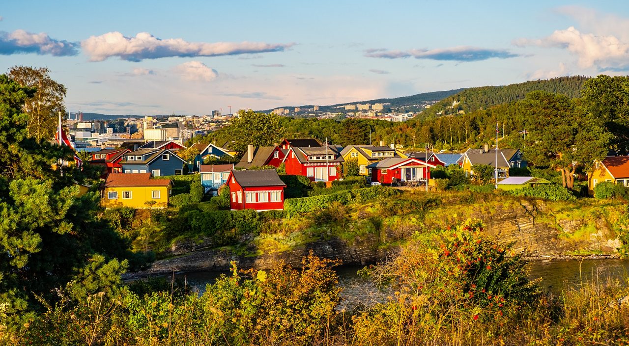 Bright red, yellow and blue houses on a green hillside above Oslo’s harbor with the modern cityscape in the background.