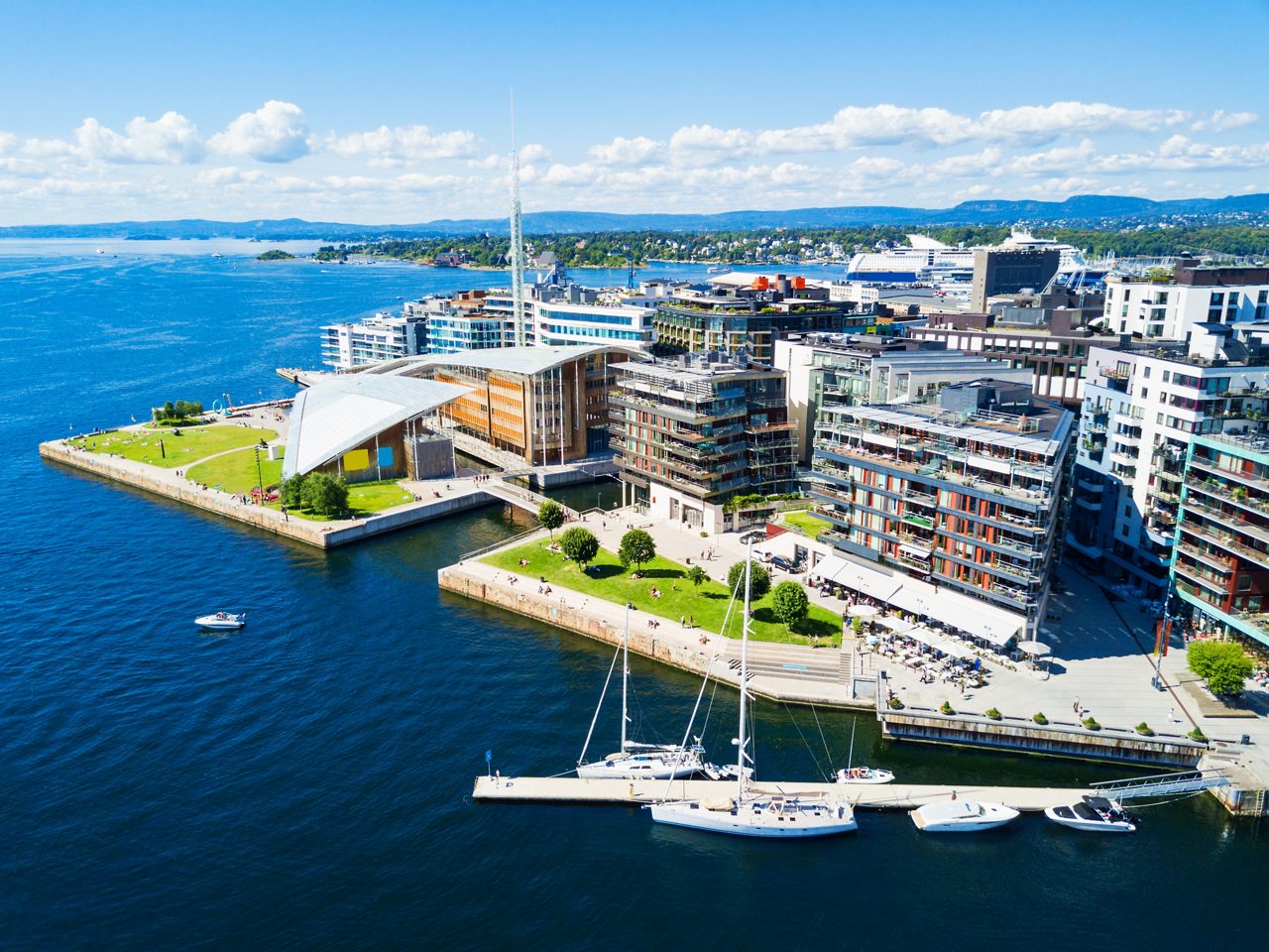  Aerial shot of a modern waterfront district featuring geometric glass buildings, grassy lawns and sailboats docked in clear blue water.