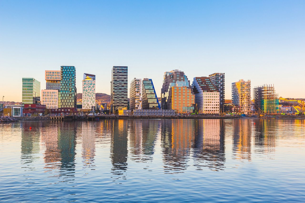 A row of modern high-rise buildings with diverse geometric designs reflects in the calm blue water of the Oslo harbor during a clear sunset.