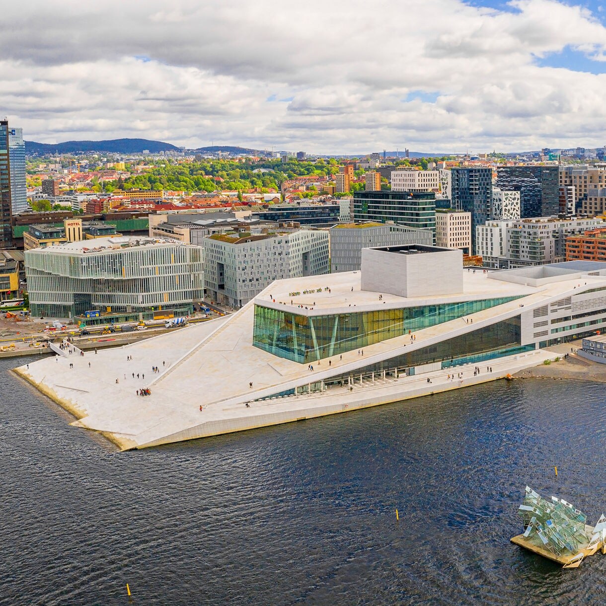 Aerial view of the Oslo Opera House with its sloping white roof beside the harbor surrounded by modern waterfront buildings and boats.