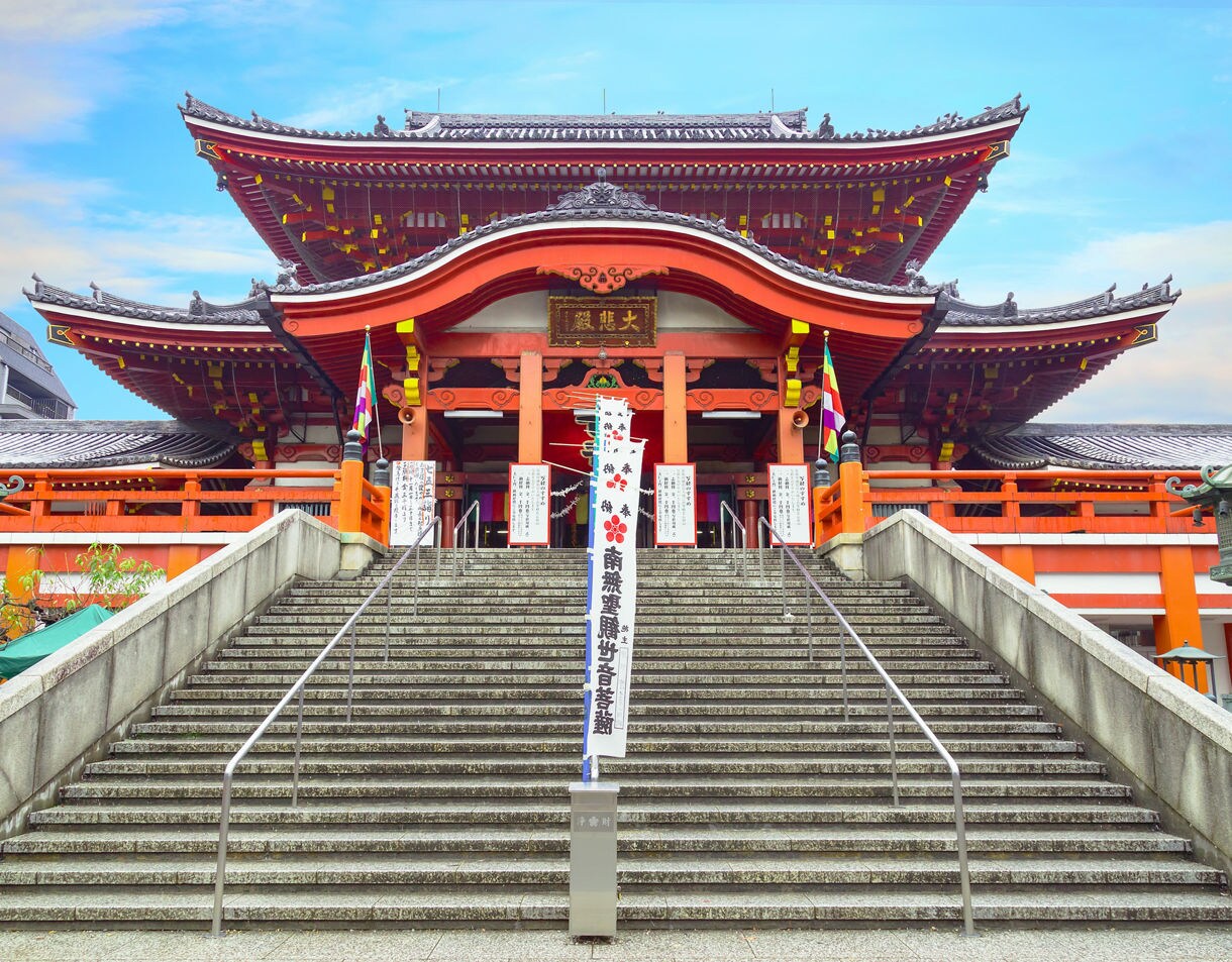 Wide stone steps lead up to a vibrant red temple in Osu with curved rooflines, hanging lanterns and banners fluttering beneath a bright blue sky.