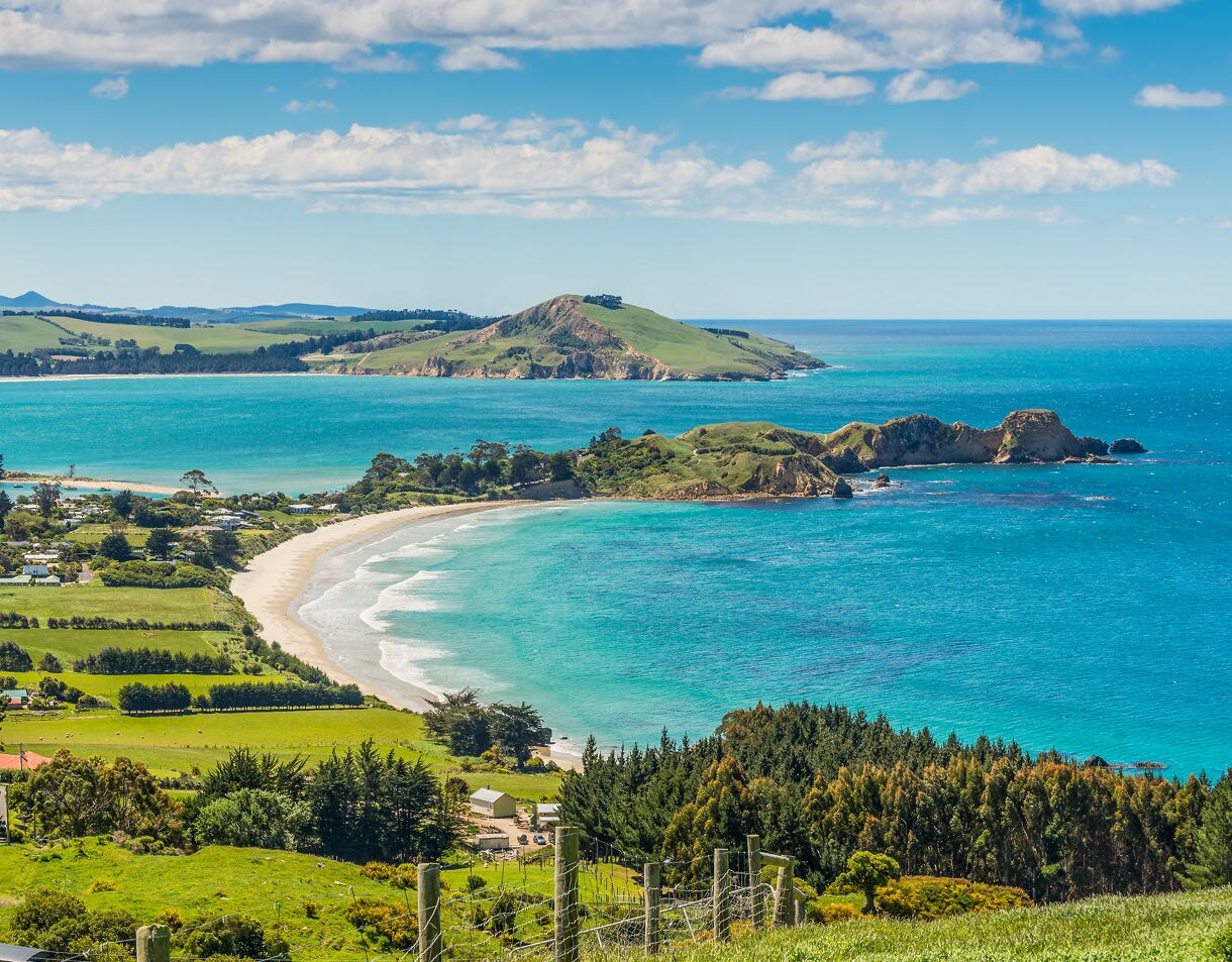 Aerial view of the Otago Peninsula’s curving beaches, green pastures and vivid blue coastal waters under a bright sky.