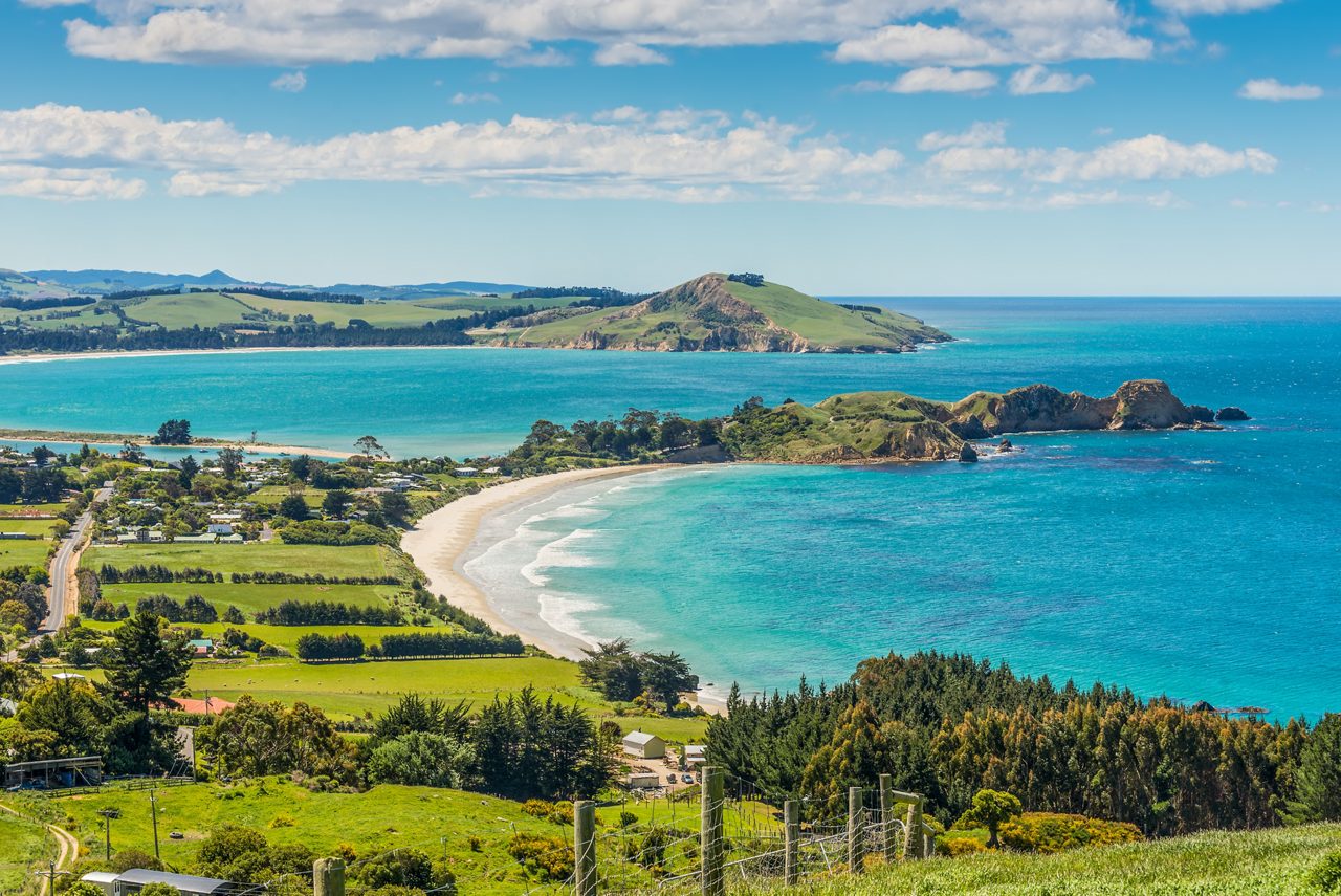 Aerial view of the Otago Peninsula’s curving beaches, green pastures and vivid blue coastal waters under a bright sky.