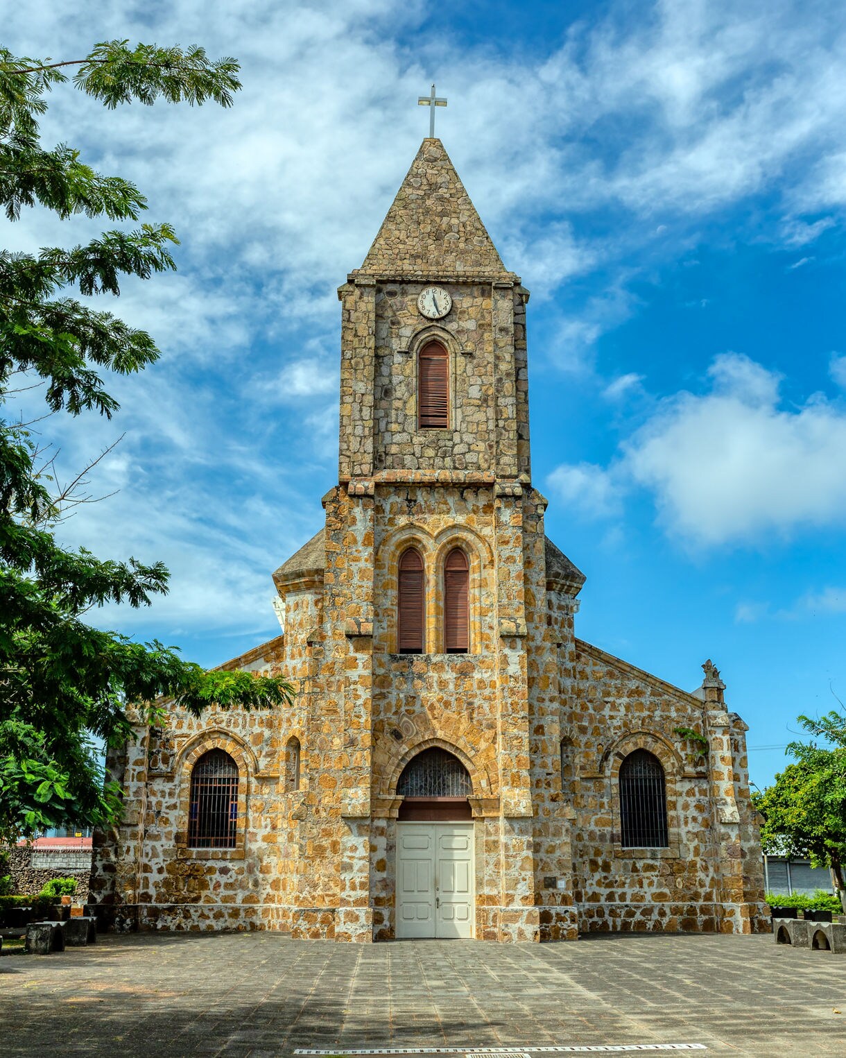 Front view of a stone church with arched windows, a clock tower and cross at the top set against a blue sky with scattered clouds.