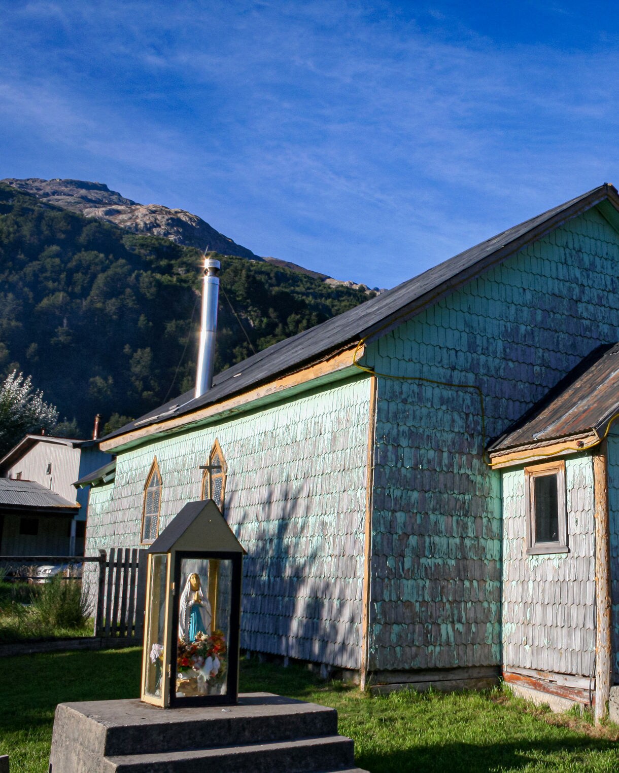 Small wooden chapel with weathered blue shingles, a simple bell tower and surrounding greenery set against forested hills and a clear sky.