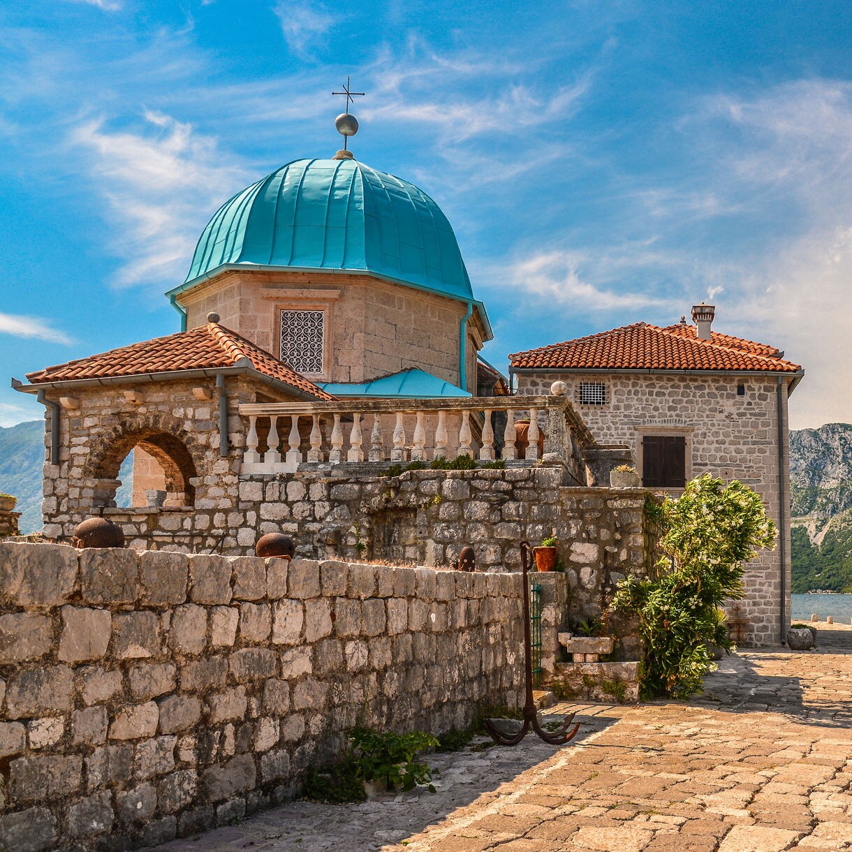 Close-up of Our Lady of the Rocks church in Montenegro, showing its turquoise dome and red-tiled roof against stone walls, with mountains and sea in the background.