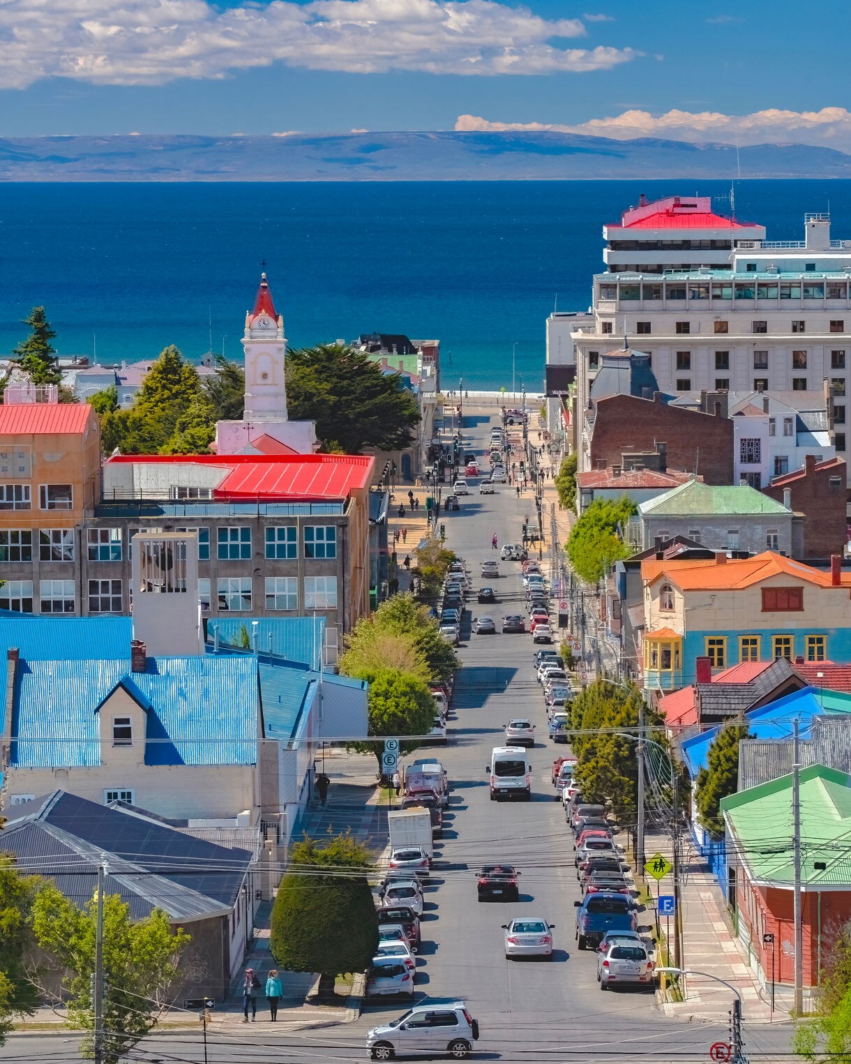 Elevated view of Punta Arenas with bright rooftops, a central street lined with cars and the blue Strait of Magellan stretching out behind the city.