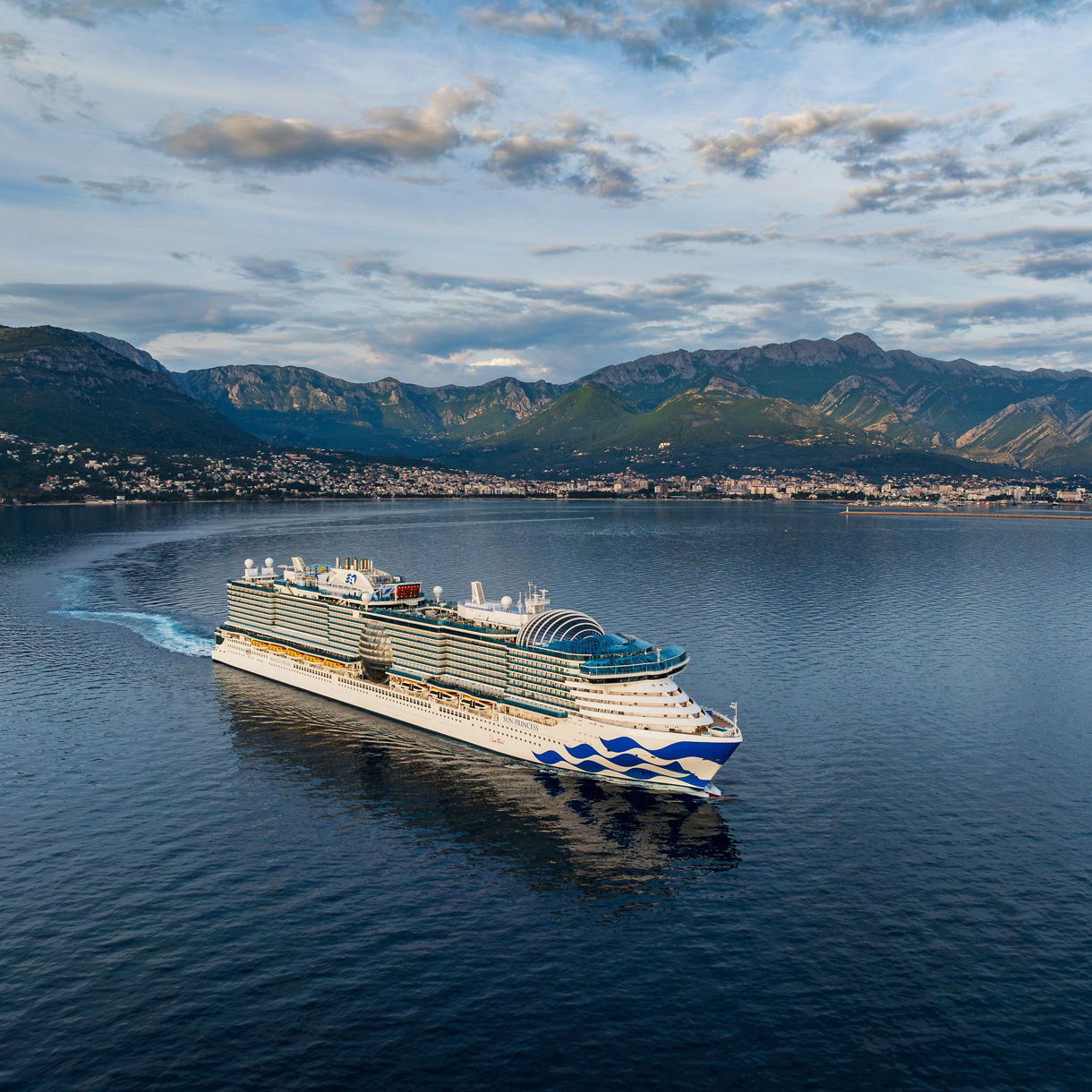 Sun Princess ship travels across calm blue waters with a scenic mountain range and coastal city in the background. The ship features multiple decks, outdoor pools, and distinctive blue wave graphics on its hull. The sky is partly cloudy, creating a tranquil and inviting atmosphere. No visible text or numbers are present on the vessel. The image captures the grandeur of maritime travel and natural beauty.