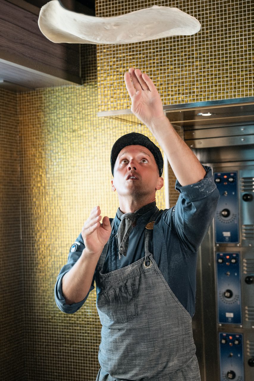 A chef in a gray apron tossing pizza dough in the air with dramatic lighting highlighting the action. 