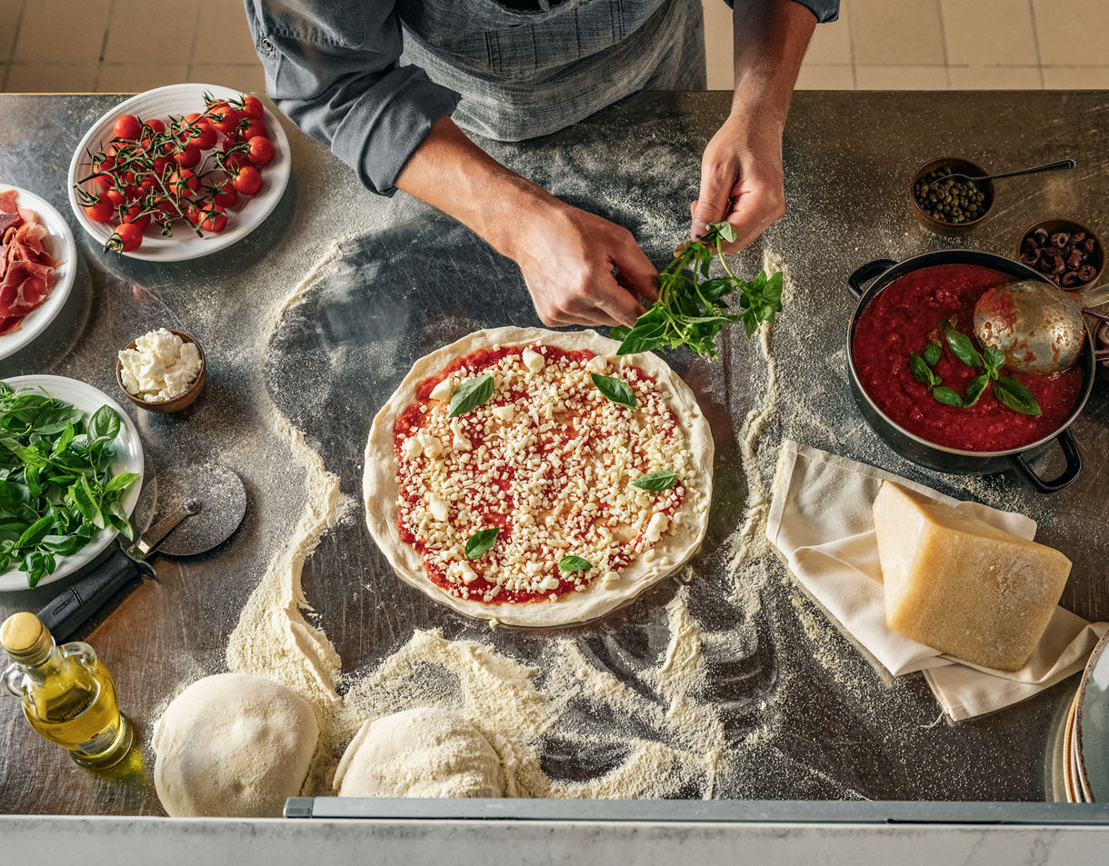 Overhead view of pizza preparation with fresh ingredients, raw dough, and cooking utensils scattered on a rustic wooden surface. 
