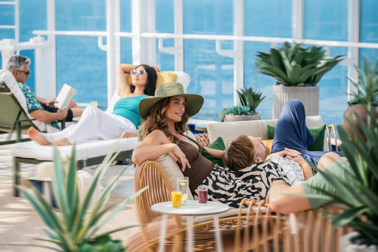     Passengers relaxing in a luxurious and sunlit lounge area on a Princess Cruises ship. A woman in a wide-brimmed hat enjoys her surroundings with a companion, while other guests lounge nearby, enjoying drinks and the peaceful ocean view. 