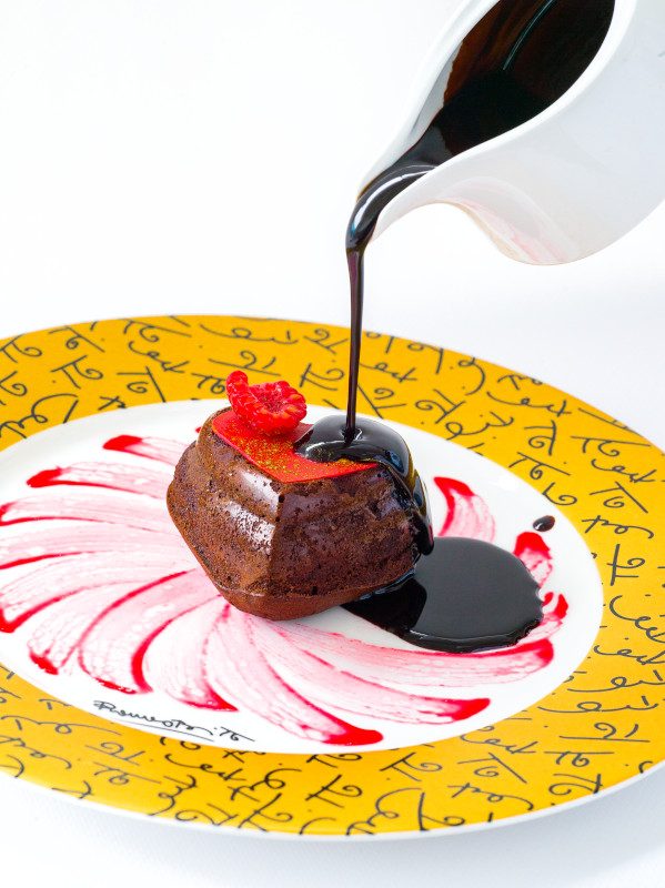 A chocolate-covered dessert being drizzled with dark sauce on a decorative plate with a yellow patterned rim and red swirl design. 