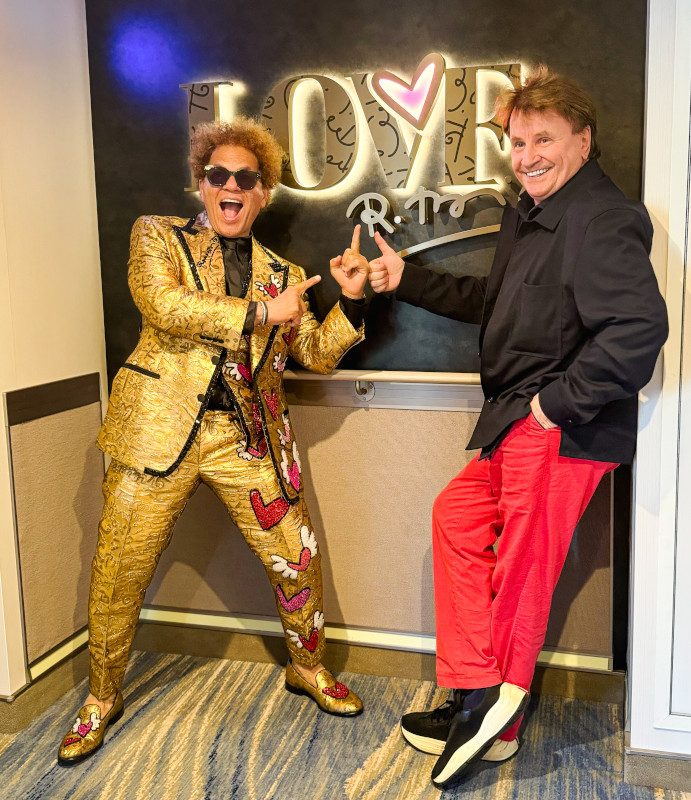 Two people posing playfully in front of a "LOVE" sign-one wearing red pants and a black jacket, the other in a gold sequined suit and sunglasses.